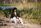 A woman sits on the riverbank looking at the camera. She is wearing a black jacket, a white shirt and white pants.