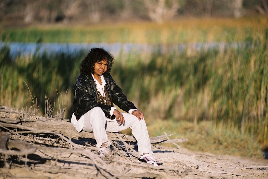 A woman is sitting on the riverbank looking at the camera. She is wearing a black jacket, a white shirt and white pants.