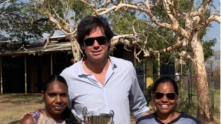 A tall man in casual clothes stands between two Aboriginal women, one holding a premiership trophy and the other two footballs.