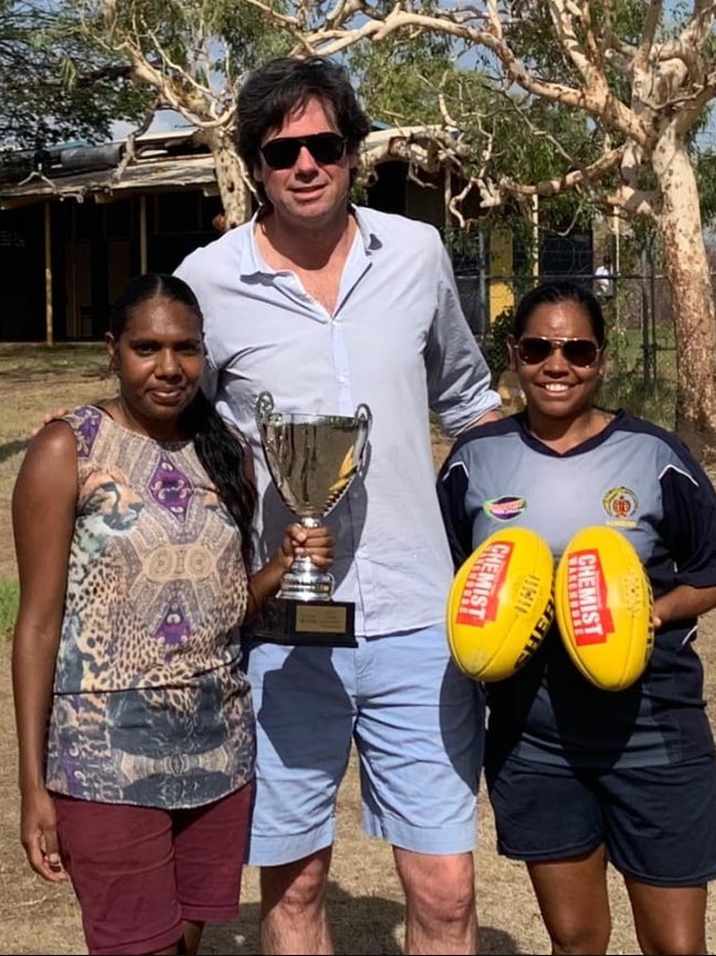 A tall man in casual clothes stands between two Aboriginal women, one holding a premiership trophy and the other two footballs.