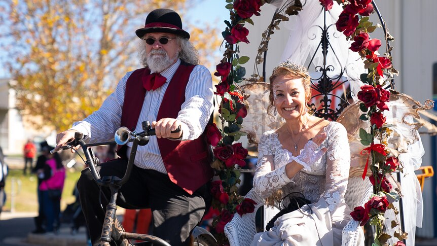 A woman dressed in white lace sits under an arch of red flowers on a wicker chair, being ridden by a man on a bicycle.