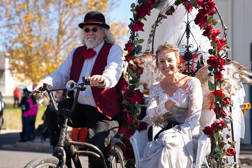 A woman dressed in white lace sits under an arch of red flowers on a wicker chair, being ridden by a man on a bicycle.