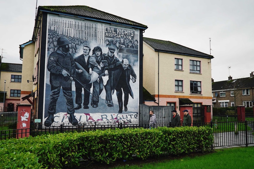 Mural on building shows protesters holding Civil Rights Association sign while soldier holds gun by people holding injured man.