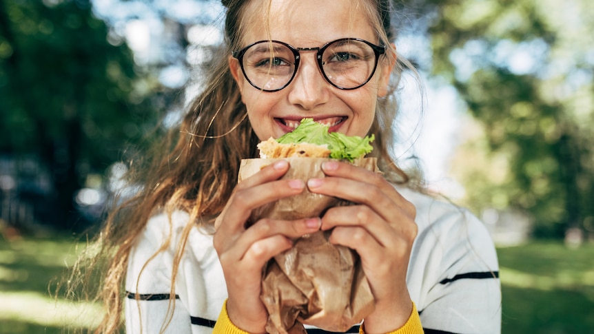 A smiling woman eats a sandwich in a park.