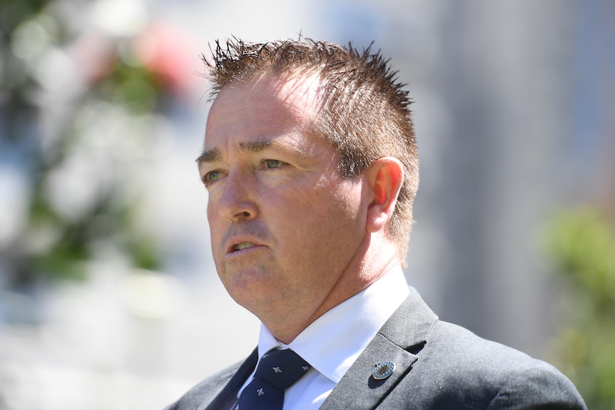 A man with short dark hair in a gray suit is standing in the sun and talking.