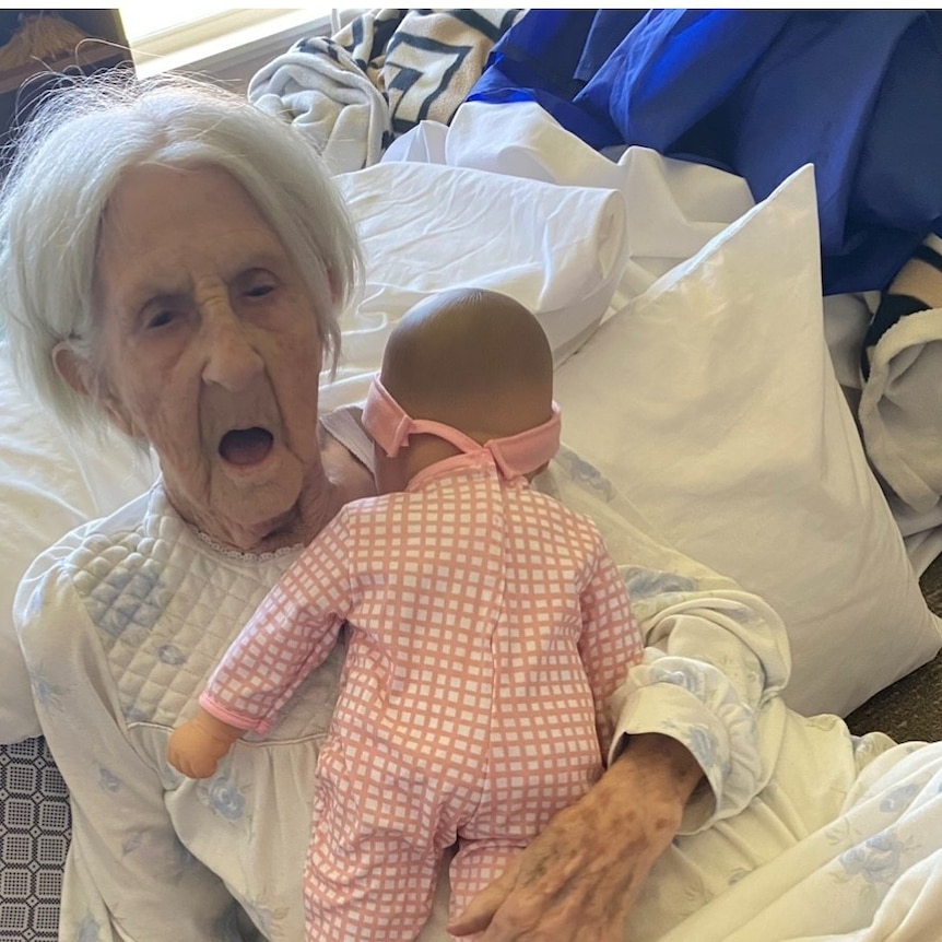 An elderly woman with white hair holds a baby doll.