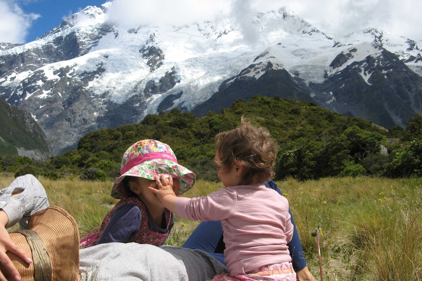 Woman lies on the grass with a hat over her head with two young children beside her. A snow-capped mountain is in the distance.