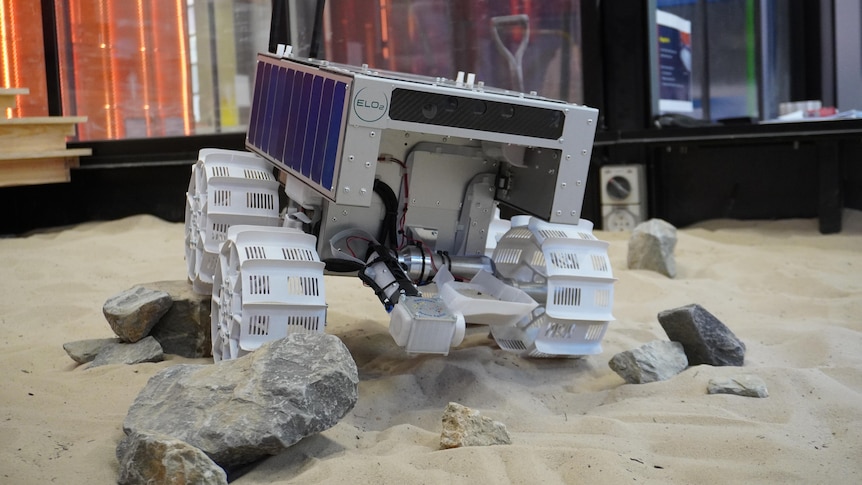 Rover on wheels sits in simulated rocky terrain with sand.
