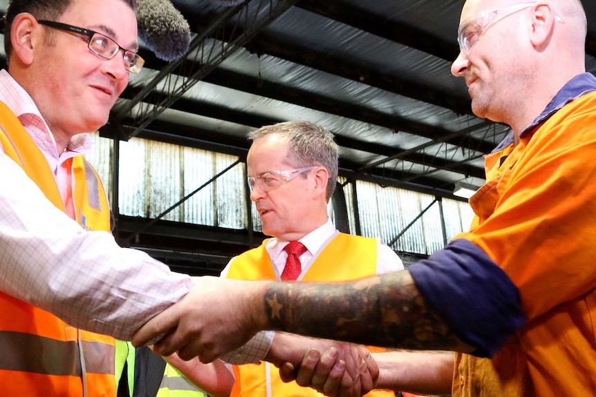 Side on view of Daniel Andrews shaking hands with an employee, foreground, with Bill Shorten looking on behind them.