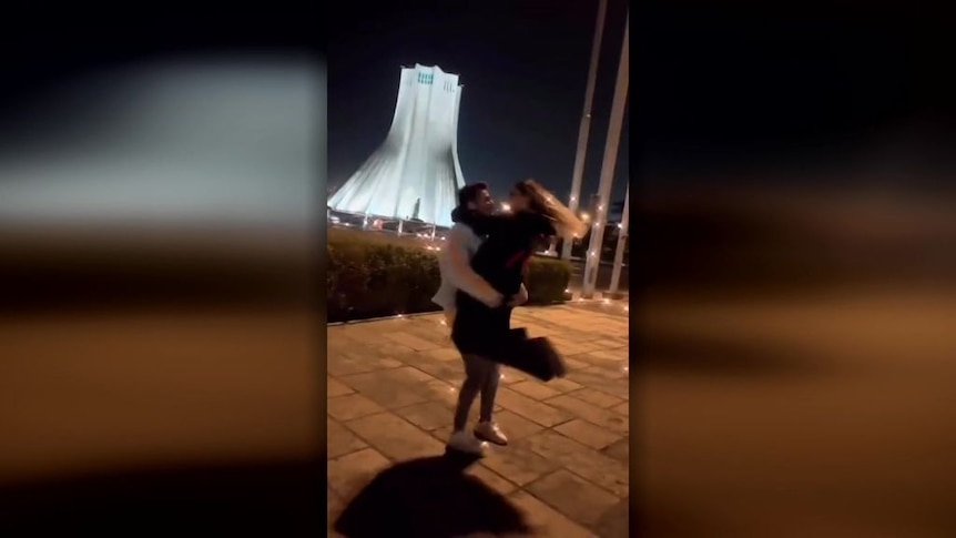 An Iranian man and woman spin around in a public square at night, with a gleaming white tower illuminated in the background.