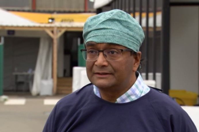 A man with glasses and green patterned surgical cap and navy medical gown stands in a car park.