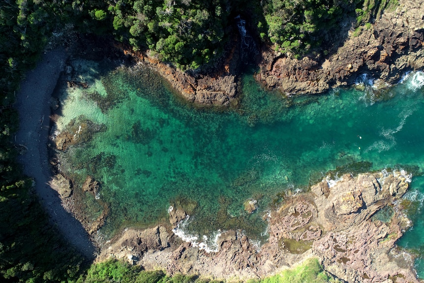 An overhead looking down photo of bright blue and green water next to a rocky coastline as seen from a drone.