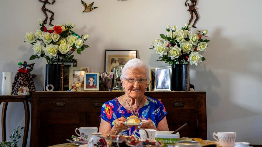 Dorothy Collishaw sits at her dining table holding a plate with a scone on it.