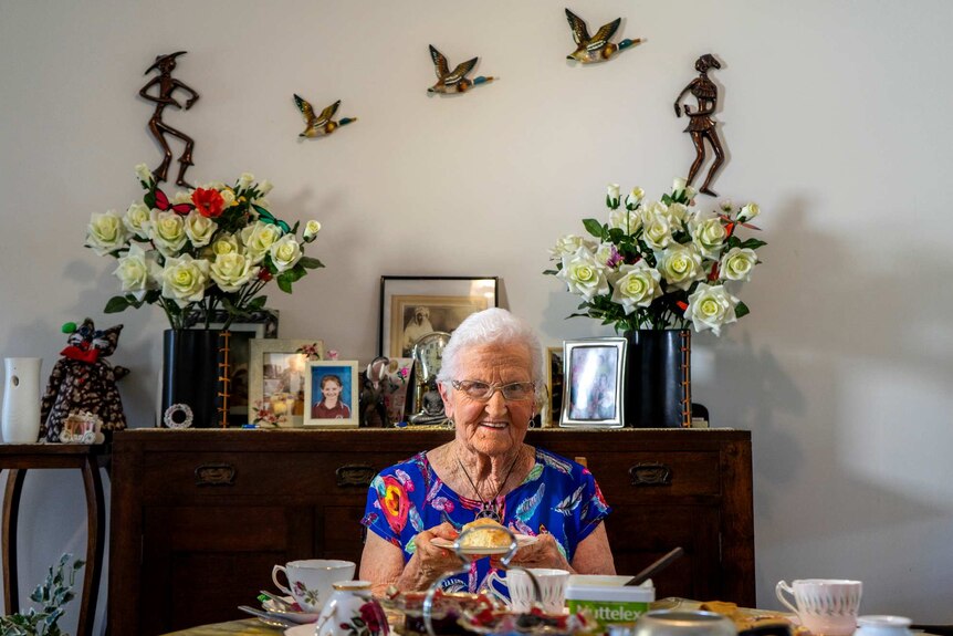 Dorothy Collishaw sits at her dining table holding a plate with a scone on it.