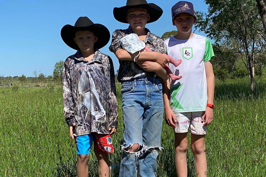 Three boys stand in a green paddock holding a small baby.