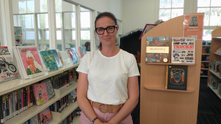 Smiling woman in her 30s, wears black-rimmed glasses, white tee, brown belt, pink pants, black hair tied, stands in a library.