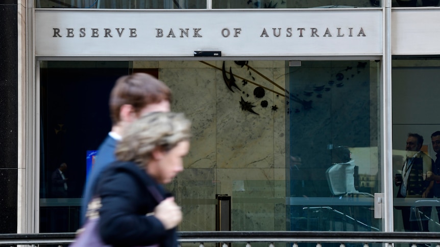 An out of focus man and woman walk past a building with glass sliding doors and a sign reading Reserve Bank of Australia.