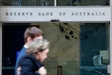 An out of focus man and woman walk past a building with glass sliding doors and a sign reading Reserve Bank of Australia.