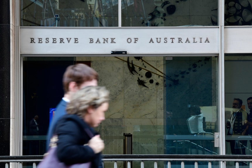 An out of focus man and woman walk past a building with glass sliding doors and a sign reading Reserve Bank of Australia.