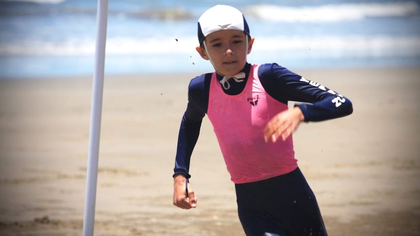 A boy in a pink and navy blue swim shirt runs on the beach.