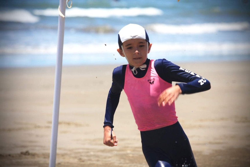 A boy in a pink and navy blue swim shirt runs on the beach.
