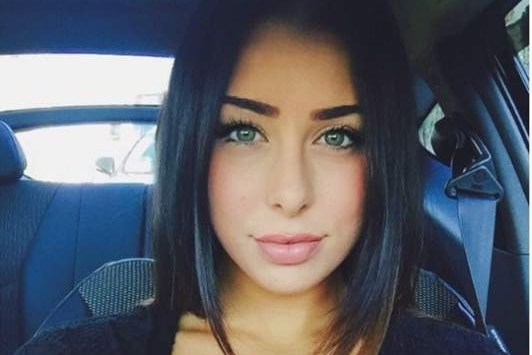 A young woman with dark hair and makeup looks at the camera as she takes a selfie in a car.