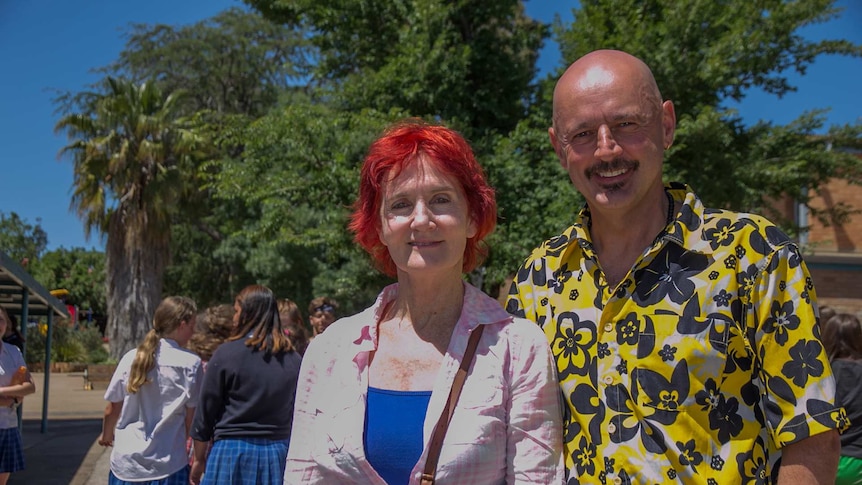 A woman with bright red hair and a man in a Hawaiian shirt smiling standing in a school playground