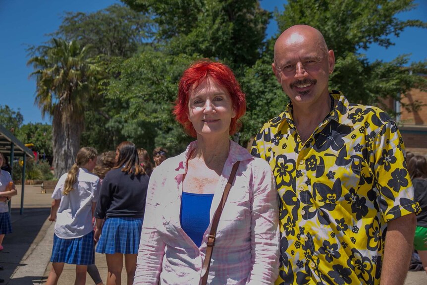 A woman with bright red hair and a man in a Hawaiian shirt smiling standing in a school playground