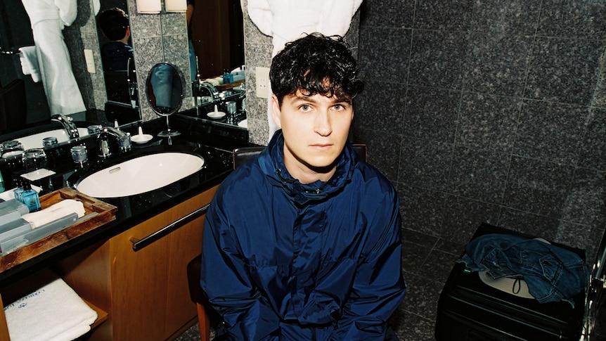 Ezra Koenig sits in a bathroom wearing a navy spray jacket.