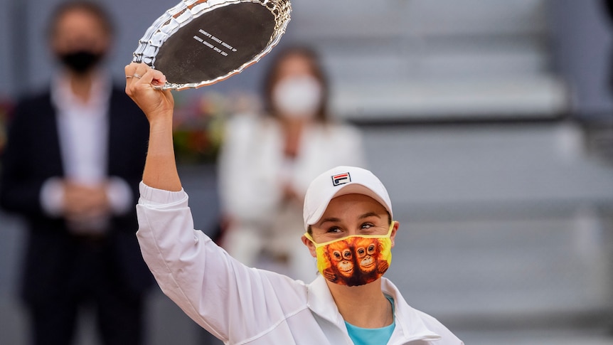 Ash Barty smiles under her mask, with two orangutans on it, while holding up a plate-style trophy in Madrid.