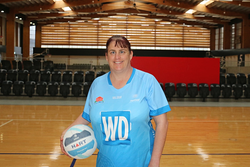 Mel stands on a netball court and smiles at the camera while wearing her blue NSW kit and holding a netball on her right hip