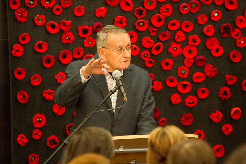An image of an older man delivering a speech, with a wall of poppies in the background.