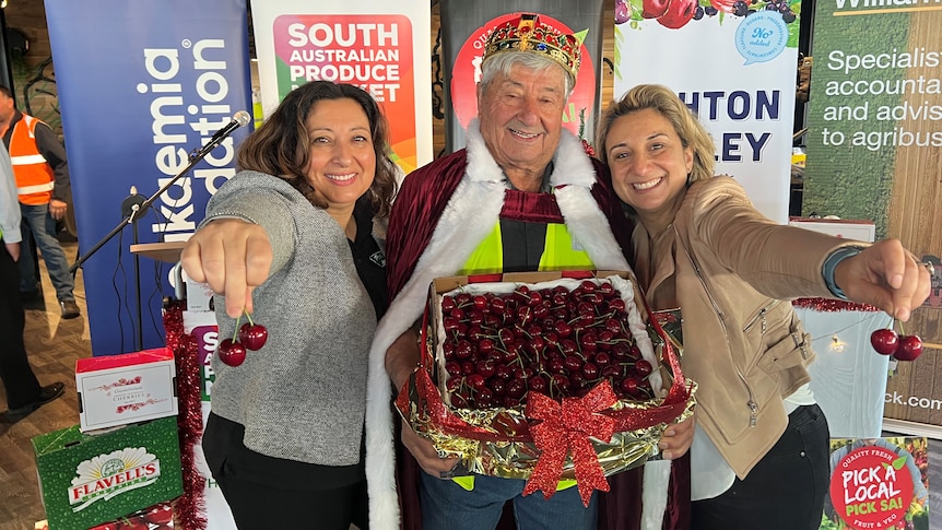 An older man with a red and gold crown and cape holding a big box of cherries with his daughters beside him