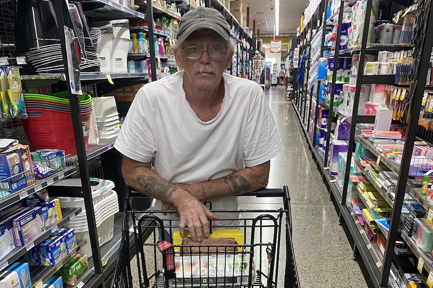 A man in his 60s in a hat and glasses, pushing a trolley inside a grocery store.