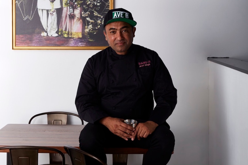 Jessi Singh smiles and wears a black chef outfit and cap, and sits on a bench inside a restaurant.