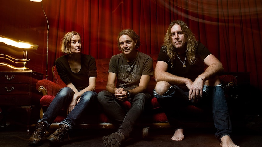 Three members of Spiderbait sitting on a red velvet couch in front of a red curtain