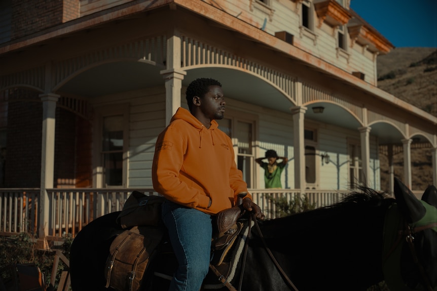 A man in an orange jumper rides a horse in front of an old house