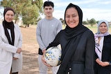 Three young Afghan women wearing hijab and a young man stand smiling in front of a brown, dusty soccer field. 