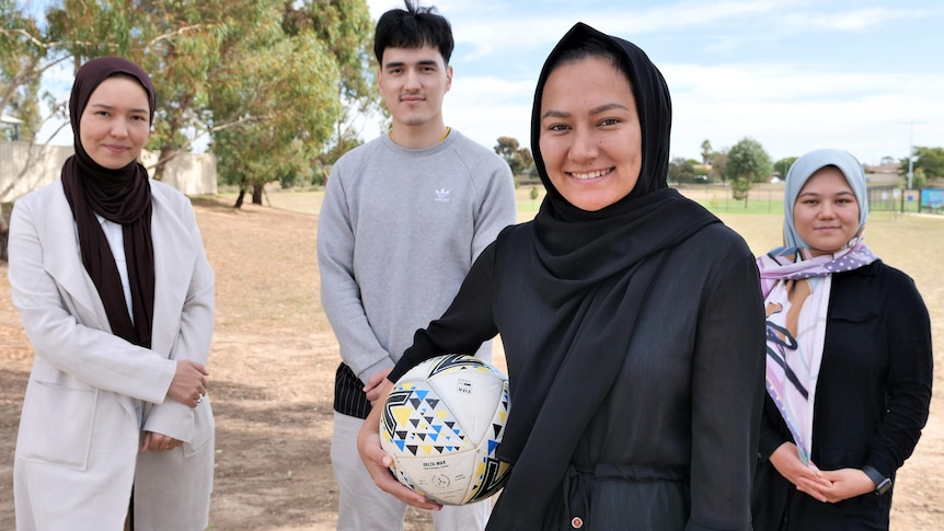 Three young Afghan women wearing hijab and a young man stand smiling in front of a brown, dusty soccer field. 