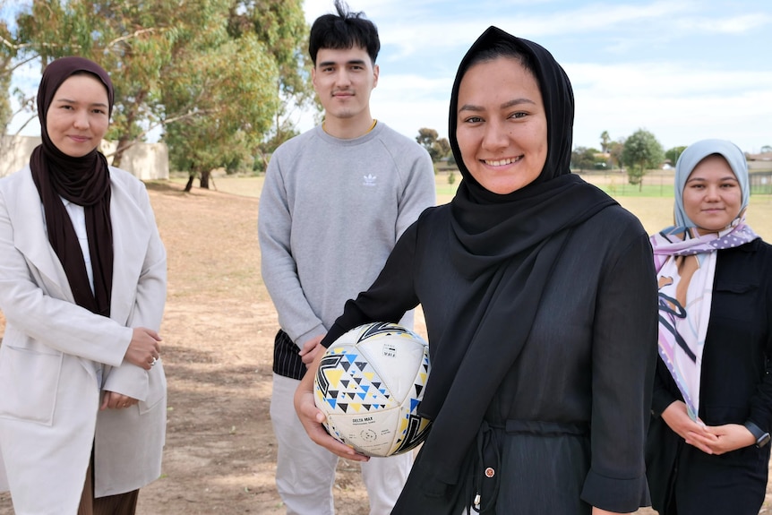 Three young Afghan women wearing hijab and a young man stand smiling in front of a brown, dusty soccer field. 