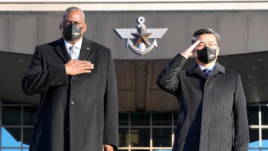 Two men wearing long black coats stand together in a building forecourt on a sunny day saluting 