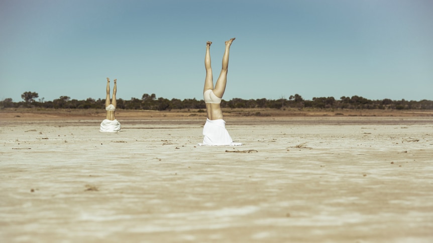 Two human figures doing handstands in desert sands with clothing covering their heads and their underwear exposed.