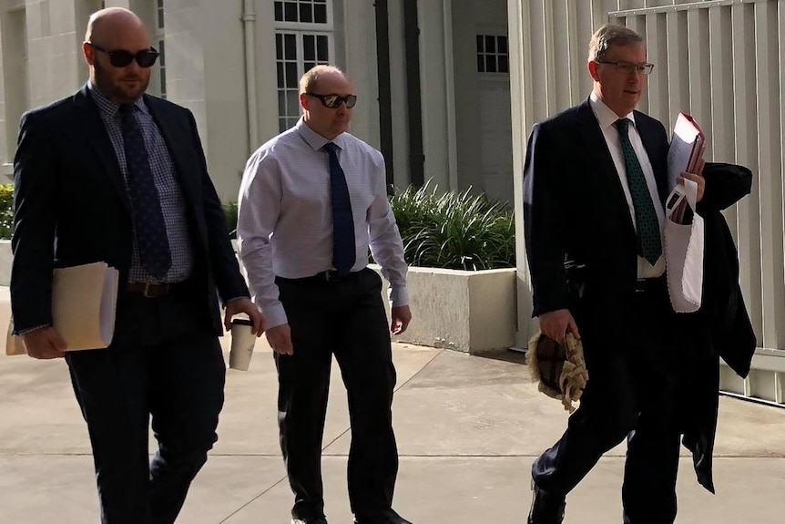 Three men in suits walk side by side along a path.