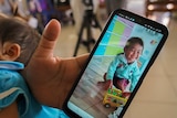 A woman holds her phone showing a photo of a smiley young toddler playing with a bus toy. A sleeping baby is in her arms