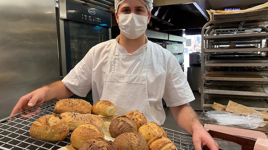 A man wearing a hairnet and facemask carrying a tray of bread loaves