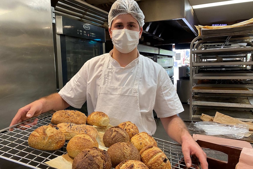 A man wearing a hairnet and facemask carrying a tray of bread loaves