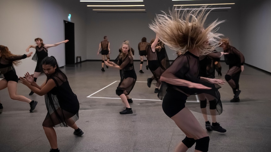In a white gallery space, a group of dancers in black chiffon outfits dance out of synchronisation.