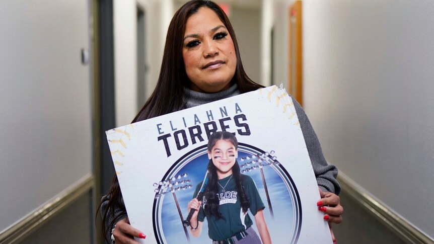 A woman holds a large poster showing a young girl in baseball gear.