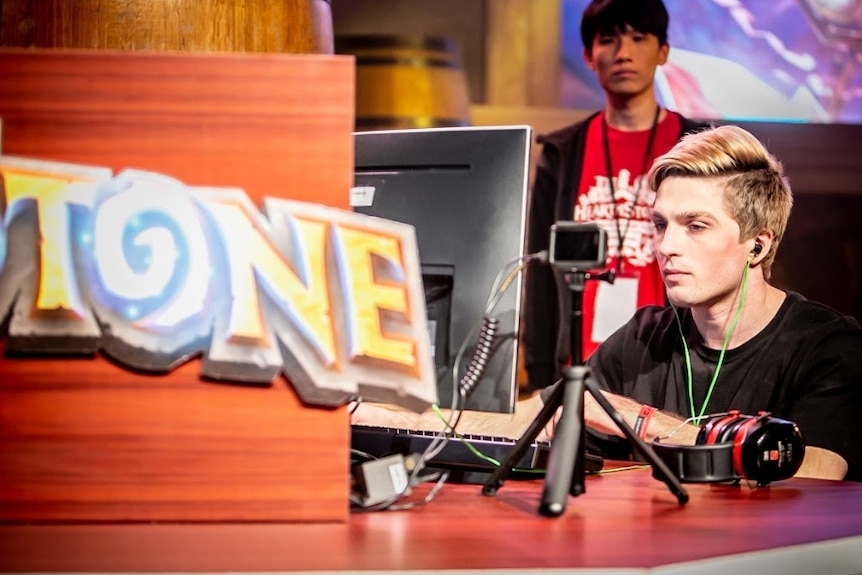 A man in a black shirt sits at a computer with headphones on in front of a sign for the Hearthstone game.