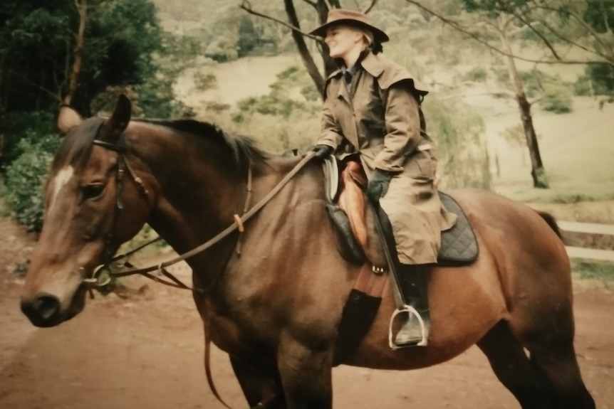 An old colored photo of a woman sitting on horse in a park, wearing a hat and a khaki trench.